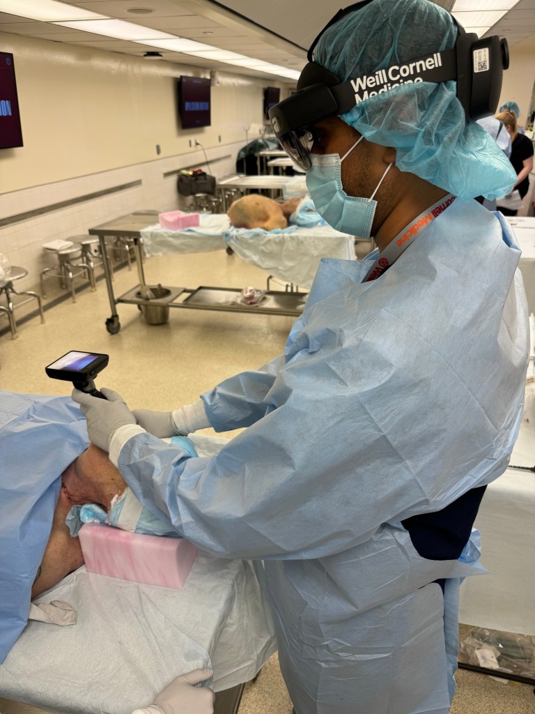 A healthcare professional wearing a blue gown and face mask engages in a procedure in a laboratory setting, utilizing a handheld device while focusing on a cadaver.