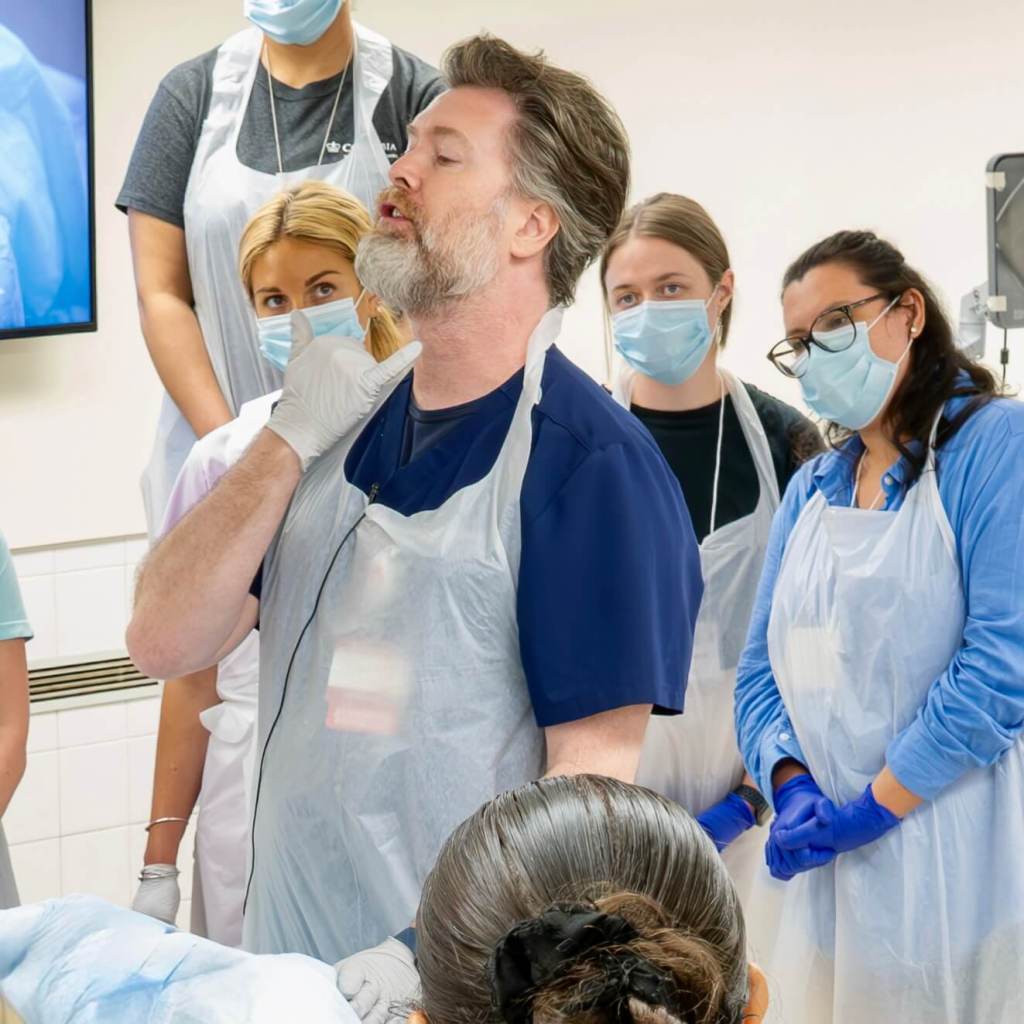 A group of healthcare professionals observing a male instructor demonstrating a procedure, all wearing masks and protective gear in an anatomy lab setting.