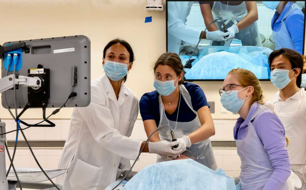 Medical professionals engaged in hands-on airway training in an anatomy lab, with a large monitor displaying a demonstration in the background.