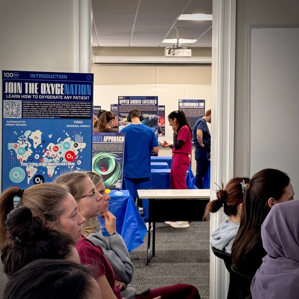 A group of healthcare professionals engaged in airway training at a workshop, with informative posters about oxygenation and airway management displayed in the background.