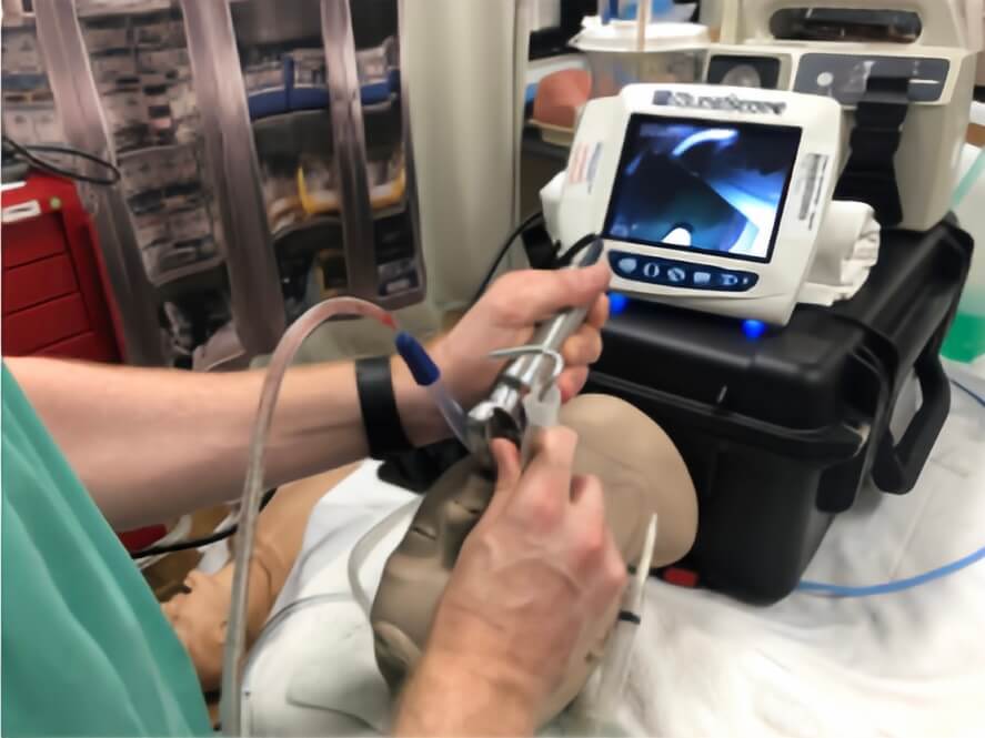 A healthcare professional using a laryngoscope and suction catheter on a training mannequin with a monitoring screen visible in the background.