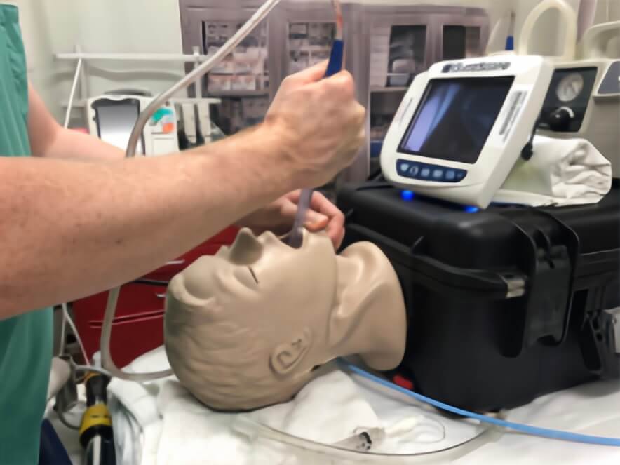 A close-up of a medical professional performing airway management training on a simulation mannequin. The individual is using a suction catheter and laryngoscope while monitoring the procedure with a device in the background.