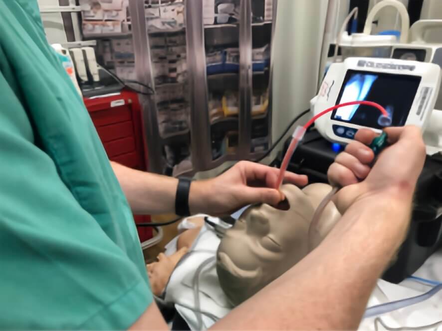 A healthcare professional performing airway management on a training mannequin using a suction catheter and video laryngoscope in a clinical setting.