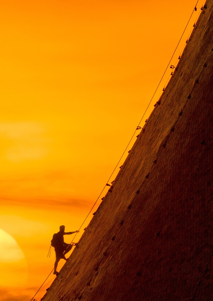 Silhouette of a climber ascending a steep surface against a vibrant orange sunset.