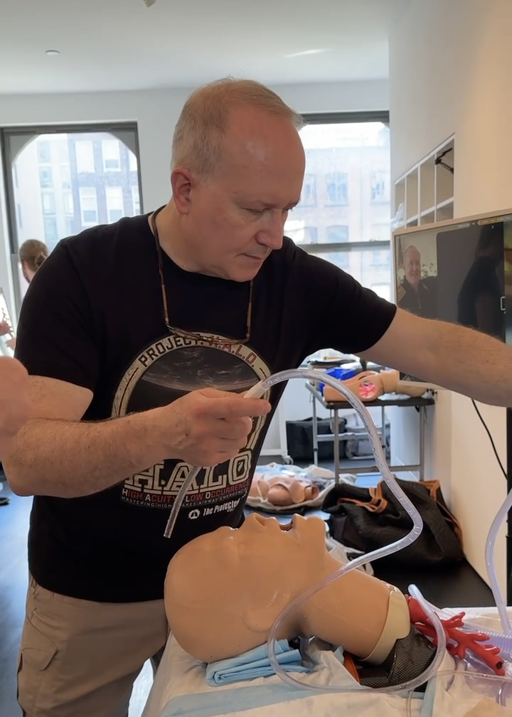 A man demonstrating medical procedures on a training mannequin while using tubing in a classroom setting.