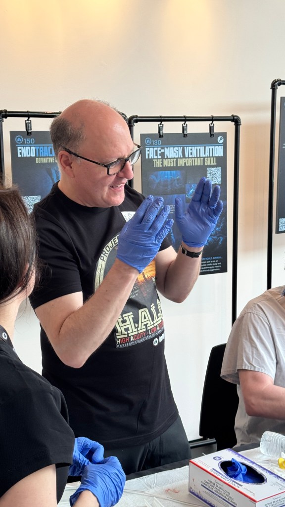 A man wearing blue gloves and glasses is demonstrating a technique, possibly related to face-mask ventilation, while two other individuals are present at a table. Educational posters are visible in the background.