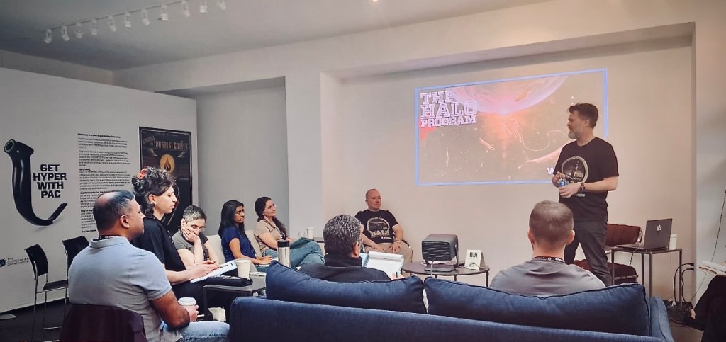 A group of people seated in a modern meeting room while a presenter standing at the front discusses a topic related to 'The Halo Program.' There is a projector screen displaying visuals relevant to the presentation.