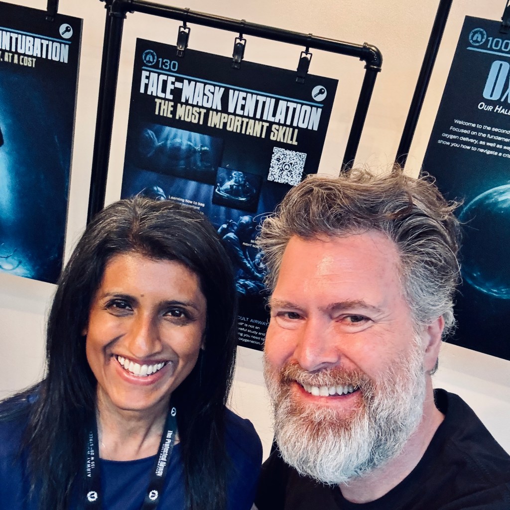 A smiling man and woman pose for a selfie in front of informational posters about face-mask ventilation and intubation at an event.